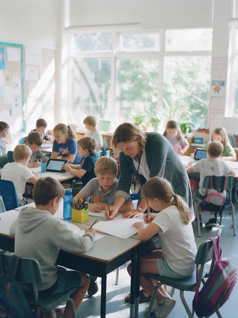 A teacher leans in to help two students at their desk while the rest of the class works in small groups
